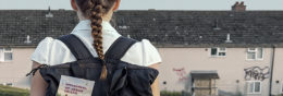 A schoolgirl with a long plait faces away from the camera towards a row of houses which have graffiti on them. The girl is wearing a backpack with a book called Memorial of Jesus' Death in the back pocket.